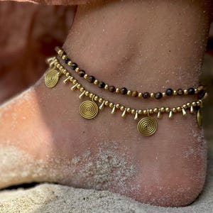 May include: Two anklets with gold-colored spiral charms and small dangling beads. The top anklet features small, dark brown beads. The anklets are worn on an ankle covered in sand, suggesting a beach setting. The jewelry is likely handmade.