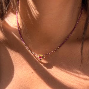 May include: A beaded necklace featuring small, faceted purple gemstone beads strung along the chain. The centre of the necklace showcases a gold-toned pendant with a rectangular red stone at its core, flanked by smaller accent stones.