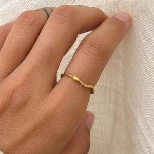 May include: A gold-colored ring with a textured, organic design. The ring is worn on a finger, showcasing its unique, nature-inspired aesthetic. The ring's surface has a subtle, irregular pattern, adding to its handcrafted appeal.