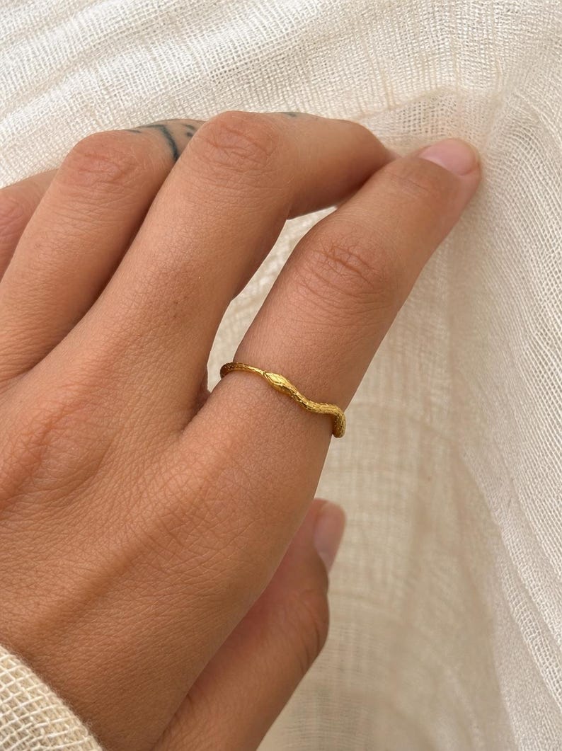 May include: A gold-colored ring with a textured, organic design. The ring is worn on a finger, showcasing its unique, nature-inspired aesthetic. The ring's surface has a subtle, irregular pattern, adding to its handcrafted appeal.