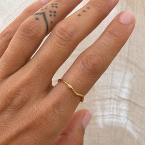 May include: A gold-colored ring with a textured, organic design is worn on a finger. The ring's surface has a wavy, branch-like pattern. The background is a soft, neutral fabric.