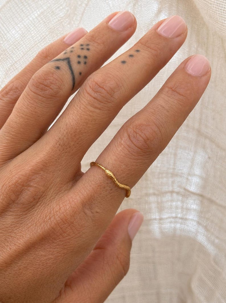 May include: A gold-colored ring with a textured, organic design is worn on a finger. The ring's surface has a wavy, branch-like pattern. The background is a soft, neutral fabric.