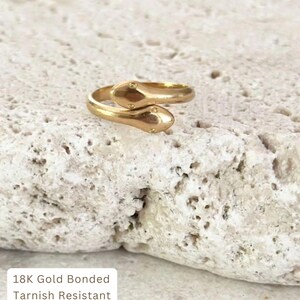 May include: A gold-colored ring in the shape of two snakes, with small dot details on the snake heads. The ring is on a textured, off-white surface. The text on the image reads: 18K Gold Bonded, Tarnish Resistant, Hypoallergenic, Water Friendly.