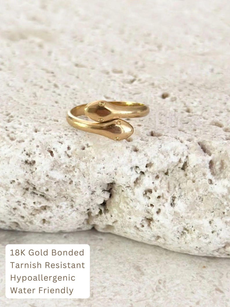 May include: A gold-colored ring in the shape of two snakes, with small dot details on the snake heads. The ring is on a textured, off-white surface. The text on the image reads: 18K Gold Bonded, Tarnish Resistant, Hypoallergenic, Water Friendly.