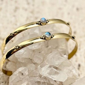 Peut inclure: Deux bracelets dorés ornés de petites pierres bleues rondes. Les bracelets sont posés sur un lit de cristaux de quartz transparents. Le design est simple, avec de petits détails décoratifs.