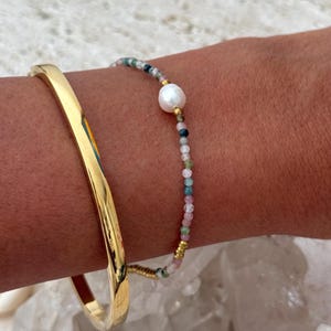 May include: A gold-coloured bangle bracelet and a beaded bracelet with a large white pearl and small colourful beads. The bracelets are on a person's wrist, with a white background.