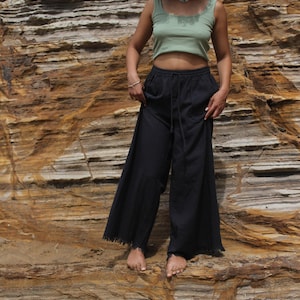 May include: A woman wearing a sage green crop top and black wide-leg pants with a drawstring waist and frayed hem. The pants are paired with a matching top. The background is a layered rock formation.