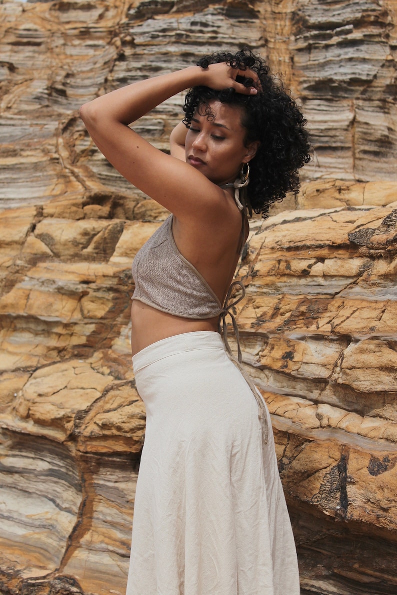 Model standing side on wearing a textured halter neck crop top which ties at the middle of back and back of neck. Light brown mushroom colour Raw cotton woven. Fitted around bust. Model wears halter top with light coloured high waisted wrap skirt.