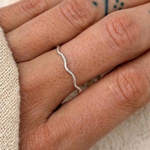 May include: A close-up of a silver ring with a wavy design. The ring is thin and appears to be made of metal. The ring is on a finger. The background is out of focus.