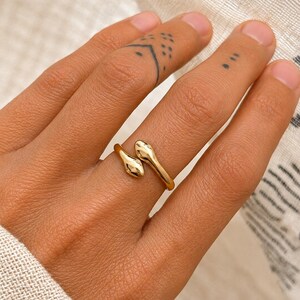 May include: A gold-colored ring in the shape of a snake, with the snake's head and tail forming the ring's ends. The ring is worn on a finger, and the skin tone is light. The background is a light-colored fabric.