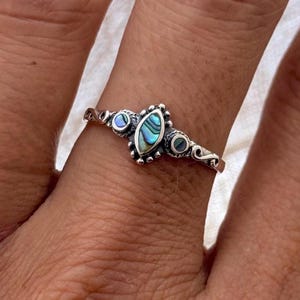May include: A silver ring with an oval abalone shell centre stone displaying iridescent blues and greens. The ring has a delicate design with small, round accent stones either side of the centre stone and decorative swirls.