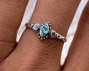 May include: A silver ring featuring an oval abalone shell center stone with iridescent blues and greens. The ring has a delicate design with small, round accent stones on either side of the center stone and decorative swirls.