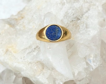 May include: A gold-colored ring with a round, deep blue stone set in the center. The ring has a smooth, polished band and a bezel setting. The stone appears to be a lapis lazuli. The ring is displayed on a white, crystalline surface.