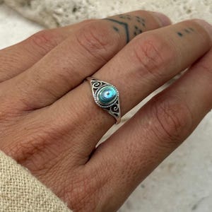 May include: A silver ring with an oval abalone shell inlay, displaying iridescent blue and green colours. The ring has a decorative filigree pattern around the shell. The ring is shown on a finger, showcasing a vintage or bohemian style.