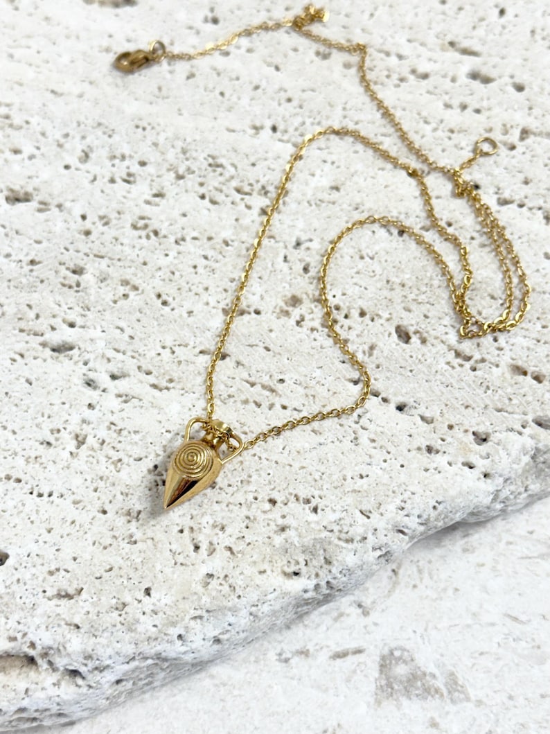 A gold-coloured necklace with an amphora-shaped pendant. The pendant has a spiral design and hangs from a fine chain. The necklace is shown on a textured, off-white surface.