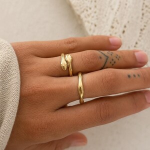 May include: Three gold rings on a hand. One ring features a snake design, with the snake's head and tail wrapping around the finger. The second ring is a simple band with a curved design. The rings are worn on a hand with tattoos.