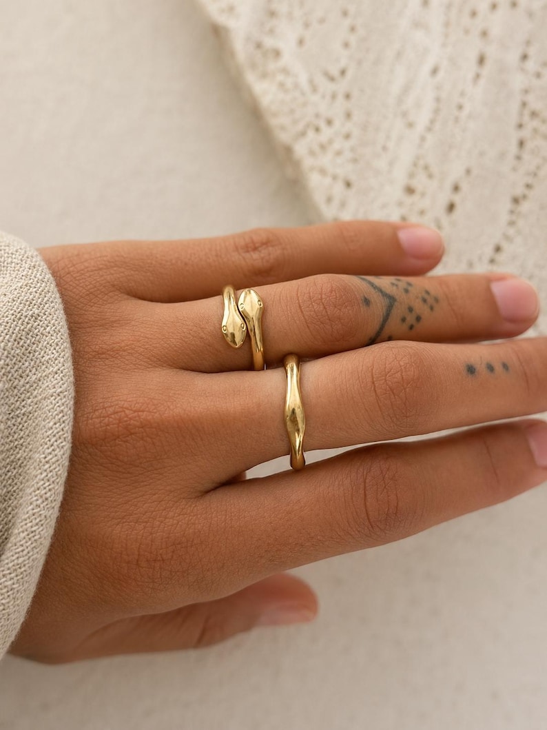 May include: Three gold rings on a hand. One ring features a snake design, with the snake's head and tail wrapping around the finger. The second ring is a simple band with a curved design. The rings are worn on a hand with tattoos.