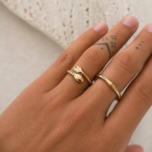 May include: Two gold rings on a hand. One ring features a snake design with two small, round details. The other ring is a simple, wavy band. The rings are set against a neutral background.