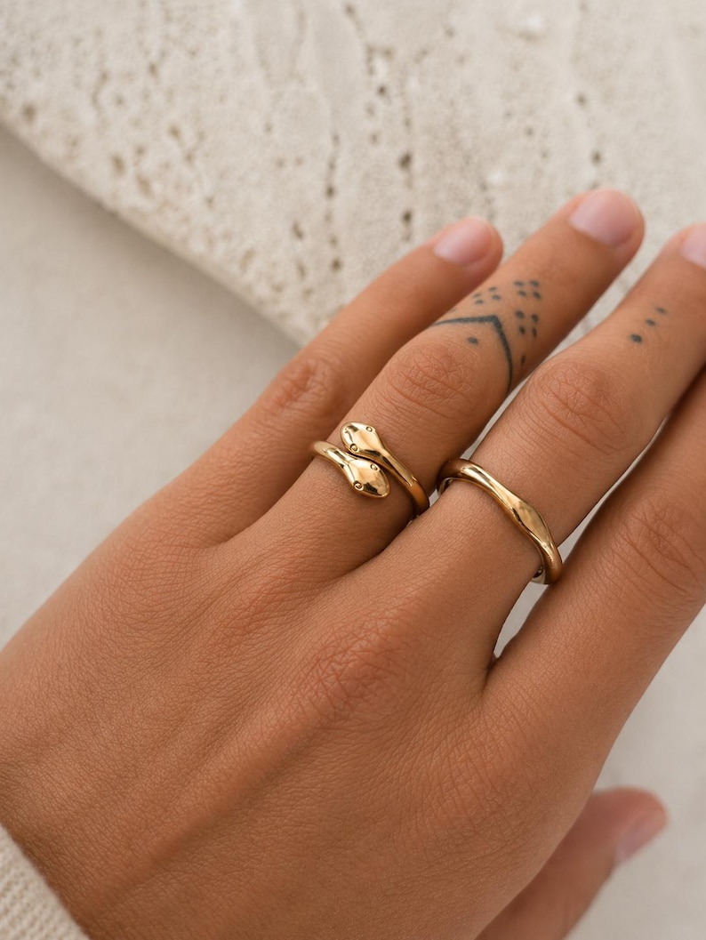 May include: Two gold rings on a hand. One ring features a snake design with two small, round details. The other ring is a simple, wavy band. The rings are set against a neutral background.