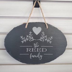 May include: A dark gray oval slate sign with a white heart and floral design. The sign reads "the REED family".