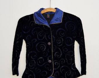 May include: A black velvet jacket featuring a tonal swirl pattern, a blue satin collar, and blue satin trim at the hem. The jacket has long sleeves, two front buttons, and gold-coloured piping along the edges. A label inside reads ro co co.