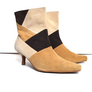 May include: A pair of suede ankle boots with a geometric color block design. The boots feature a pointed toe and a small, brown heel. The color scheme includes beige, tan, and black panels.