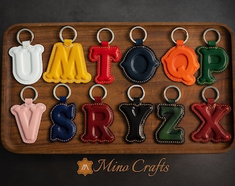 May include: A set of 12 leather keychains, each featuring a different letter of the alphabet. The keychains are in various colors, including white, yellow, red, blue, orange, green, and black. The keychains are arranged in two rows on a wooden tray. The tray has the text "Mino Crafts" printed on it.