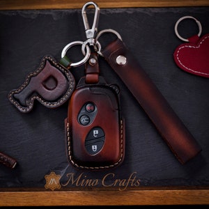 May include: A brown leather key fob with a key chain and a leather strap. The key fob has a brown leather letter "P" attached to it. The key fob is on a black surface with the text "Mino Crafts" visible.
