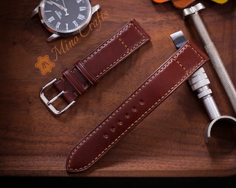 May include: Two brown leather watch straps with contrasting stitching. The straps are laid out on a wooden surface with a silver watch and leather working tools.
