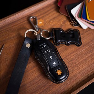 May include: Black leather key fob with a brown stitching detail. The fob has a small metal loop and a black leather strap. The fob is designed to fit a car key with four buttons. The fob has a small leather tag with the letter "M" on it. The text "Mino Crafts" is visible in the background.