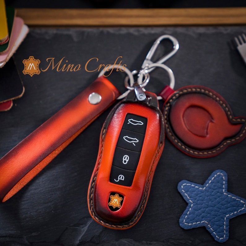 May include: A red leather key fob with a silver key ring and a matching leather strap. The key fob has a logo of a letter "M" in the centre. The key fob is designed to fit a car key.