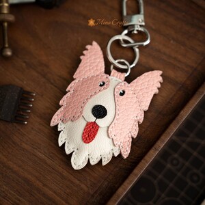 May include: A leather dog-shaped keychain with a pink and white design. The dog has a black nose, red tongue, and a silver keyring. The keychain is on a wooden surface.
