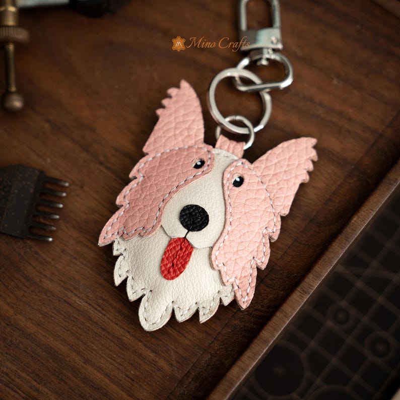 May include: A leather dog-shaped keychain with a pink and white design. The dog has a black nose, red tongue, and a silver keyring. The keychain is on a wooden surface.
