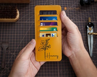 May include: Mustard yellow leather card holder with six card slots. The front features a floral design and the word "Harmony" in black. The card holder is stitched with a contrasting light-colored thread.