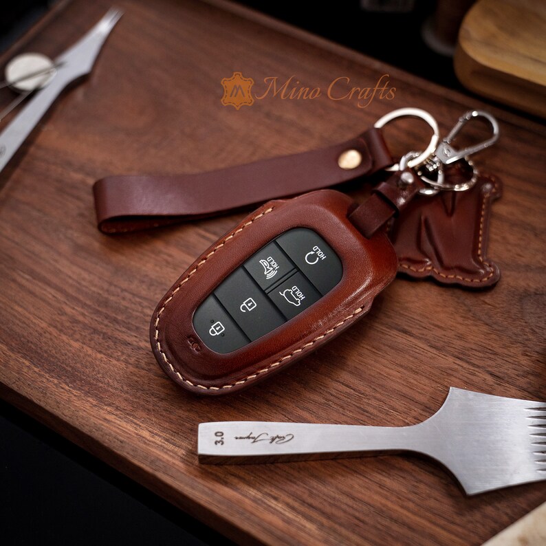 May include: A brown leather key fob with a key chain and a leather strap. The key fob has a stitched edge and a metal key ring. The key fob is on a wooden surface.