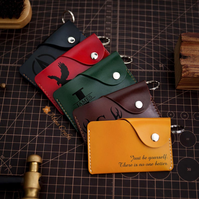 May include: Five leather keychains with different designs and engravings. The keychains are in various colors, including black, red, green, brown, and yellow. The engravings include a compass, a bird, the name "JAMES", a deer, and the quote "Just be yourself. There is no one better."