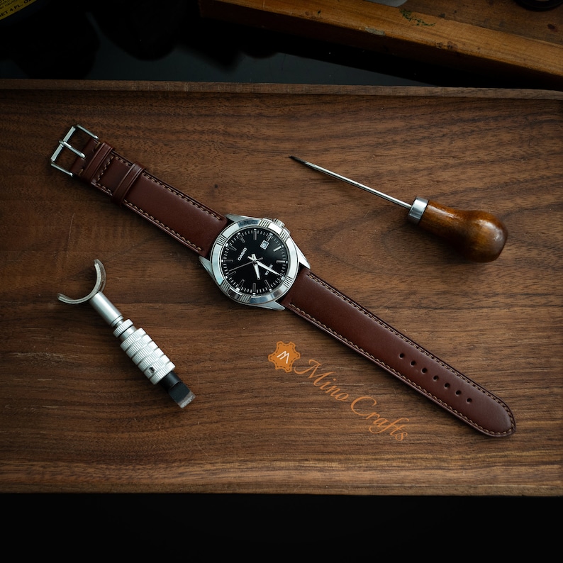 May include: A brown leather watch strap with stitching detail on a wooden surface. The strap is attached to a silver watch with a black face. The watch is surrounded by leather working tools.