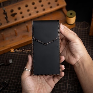 May include: Black leather card holder with a flap closure and contrasting brown stitching. The card holder is rectangular and features a stitched border. The Mino Crafts logo is visible in the lower left corner.