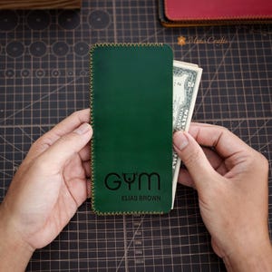 May include: Green leather phone case with a gold stitched border. The case has a black logo that says "GYM" with a dumbbell icon and "ELIAS BROWN" underneath.