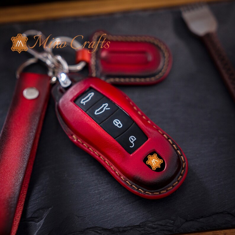 May include: Red leather key fob with stitching and a small logo. The key fob has four buttons and a metal ring.
