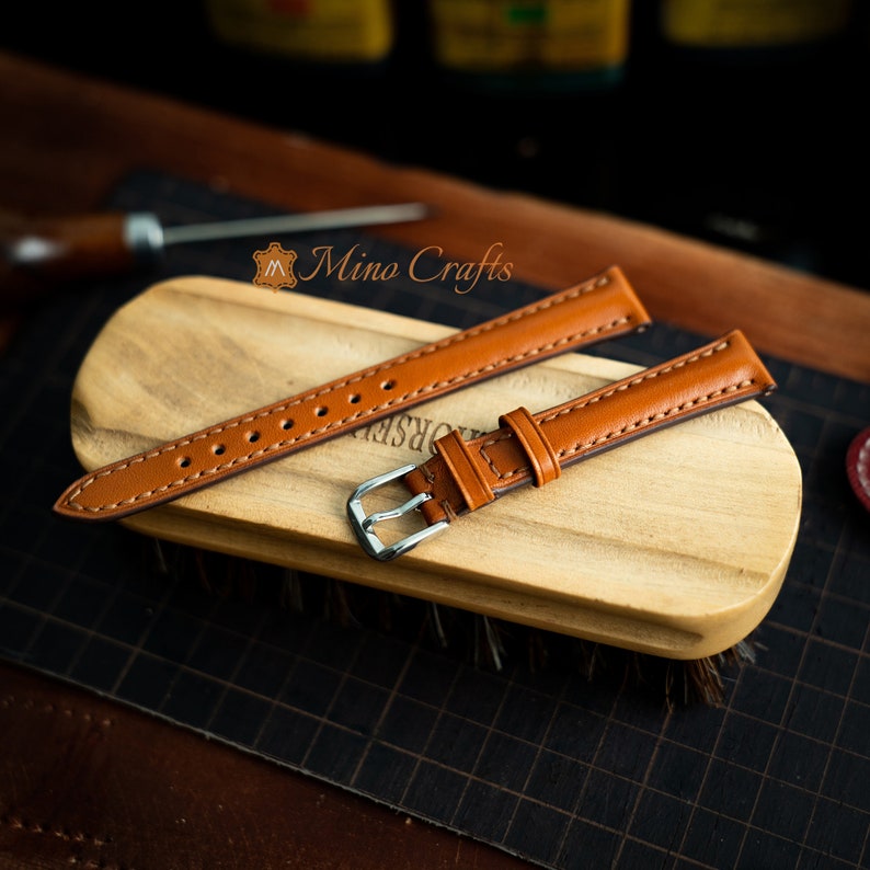 May include: Two brown leather watch straps with silver buckles. The straps are laid out on a wooden surface. Mino Crafts is printed on the surface.