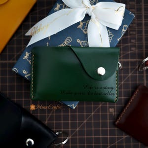 May include: A green leather wallet with a flap closure and a silver snap. The wallet has a quote engraved on the front: "Life is a story. Make yours the best seller."