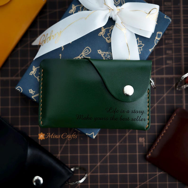 May include: A green leather wallet with a flap closure and a silver snap. The wallet has a quote engraved on the front: "Life is a story. Make yours the best seller."