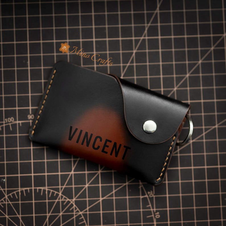 May include: A brown leather key holder with a black flap and a silver snap closure. The key holder has the name "VINCENT" embossed on the front.