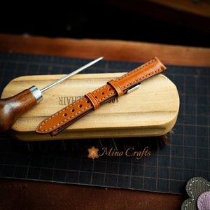 May include: A brown leather watch strap with white stitching and a silver buckle. The strap is lying on a wooden surface.