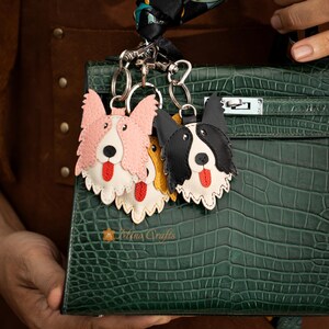 May include: Three leather dog-shaped keychains in pink, black and white, and yellow, each with a red tongue. The keychains are attached to a green handbag. The Mina Crafts logo is visible on the handbag.