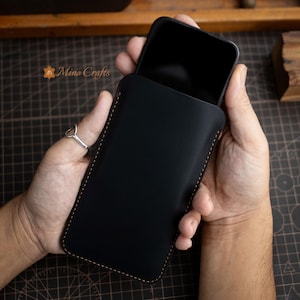 May include: A black leather phone sleeve with a contrasting tan stitch detail. The phone case is held in a person's hands. The brand name "Mino Crafts" is visible in the upper left corner.