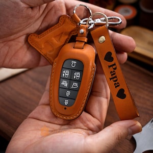 May include: A brown leather key fob with a key chain and a leather strap with the word "Papa" and a heart design. The key fob has a grey button with the word "Hold" and a car icon.