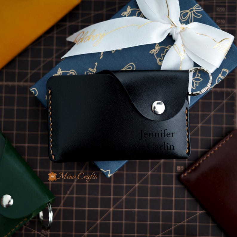 May include: Black leather key chain with a flap and a snap closure. The keychain has a silver metal ring and is personalized with the name "Jennifer Carlin".