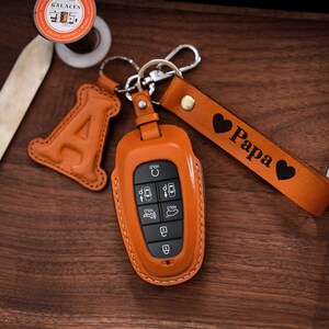 May include: A brown leather key fob with a key chain attached. The key fob has a leather strap with the word "Papa" engraved on it. The key fob is decorated with two hearts.